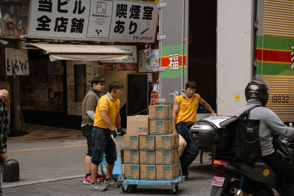 荷物を台車に積む作業着の人たちと、そばを通るバイク