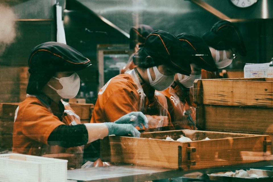 食品の加工場で、作業帽とマスクを着けた人たちが、並んで箱詰めをしている
